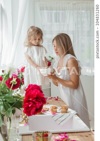 A little blonde girl with her mom on a kitchen countertop decorated with peonies. The concept of the relationship between mother and daughter. Spring atmosphere. 104831260