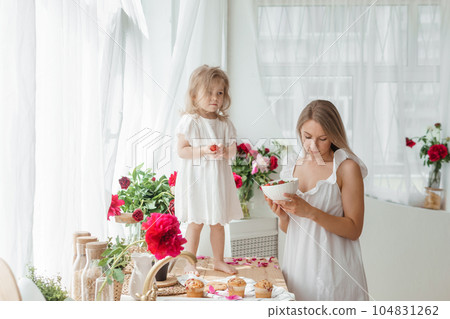 A little blonde girl with her mom on a kitchen countertop decorated with peonies. The concept of the relationship between mother and daughter. Spring atmosphere. A little blonde girl with her mom on a kitchen countertop decorated with peonies. The concept of the relationship between mother and daughter. Spring atmosphere. 104831262