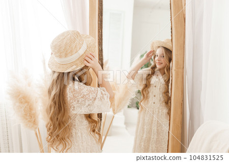 A beautiful teenage girl with long hair measures a straw hat in front of a mirror. Self-admiration of a blonde. A beautiful teenage girl with long hair measures a straw hat in front of a mirror. Self-admiration of a blonde. 104831525