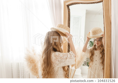 A beautiful teenage girl with long hair measures a straw hat in front of a mirror. Self-admiration of a blonde. A beautiful teenage girl with long hair measures a straw hat in front of a mirror. Self-admiration of a blonde. 104831526