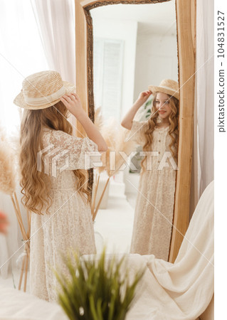 A beautiful teenage girl with long hair measures a straw hat in front of a mirror. Self-admiration of a blonde. 104831527