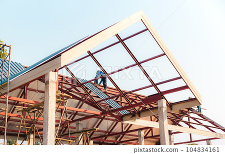 Roofer working in special protective work wear gloves, using air or pneumatic nail gun installing asphalt shingle on top of the new roof. 104834161