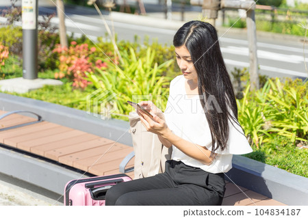 一位年輕女子坐在長凳上,在手提箱旁邊操作智能手機 一位年輕女子坐在長凳上,在手提箱旁邊操作智能手機 104834187
