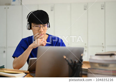 Concentrated male medical students wearing headphone and reading book in library. Education and Technology concept Concentrated male medical students wearing headphone and reading book in library. Education and Technology concept 104835216