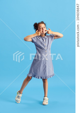 Full-length portrait of little girl, child with curly hair and cute hairstyle posing with grimacing face against blue studio background 104836647