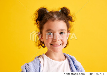 Close-up portrait of beautiful little girl, child with curly hair posing, looking at camera, smiling against yellow studio background 104836711