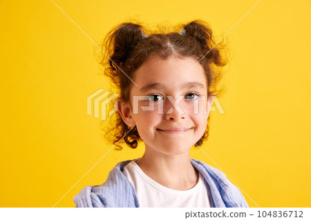 Close-up portrait of beautiful little girl, child with curly hair posing, looking at camera, smiling against yellow studio background 104836712