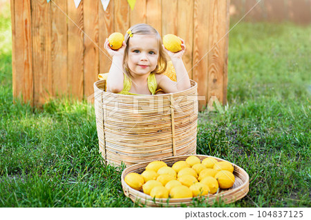 Little child girl with lemons at lemonade stand in park. Portrait of funny baby in basket with fruits. vitamins and healthy food. Useful citrus fruits as prevention of viral and respiratory diseases. 104837125