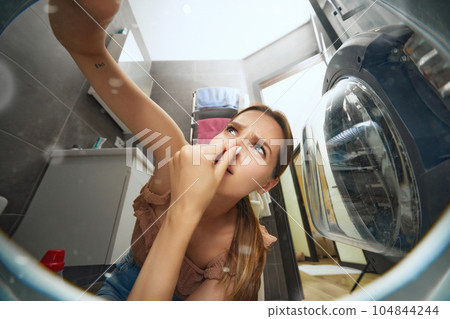 Young woman doing domestic chores, putting dirty clothes into washer. Bad smell. View from inside of washing machine 104844244