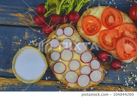 Spread butter on bread with sliced tomatoes and radishes. Fresh snack on natural wooden background. Flat design. Top view Spread butter on bread with sliced tomatoes and radishes. Fresh snack on natural wooden background. Flat design. Top view 104844260