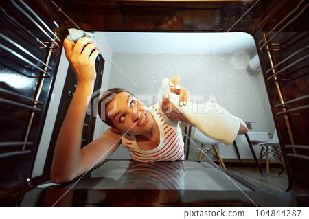 Young woman emotionally cleaning oven with effective cleaning products. After cooking kitchen clean up. View from oven Young woman emotionally cleaning oven with effective cleaning products. After cooking kitchen clean up. View from oven 104844287