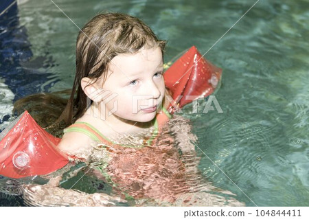 Small blond girl with armband floats in swimming pool. Child learns to swimm. 104844411