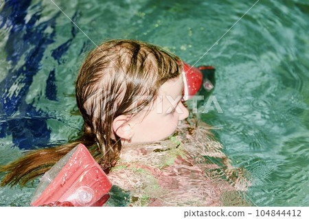 Small blond girl with armband floats in swimming pool. Child learns to swimm. Small blond girl with armband floats in swimming pool. Child learns to swimm. 104844412