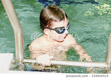 Boy in the indoor public pool. Portrait of child with swimmng goggles. Boy goes up on ladder. 104844424