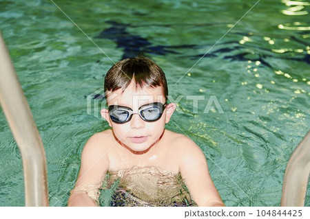 Boy in the indoor public pool. Portrait of child with swimmng goggles. Boy goes up on ladder. 104844425