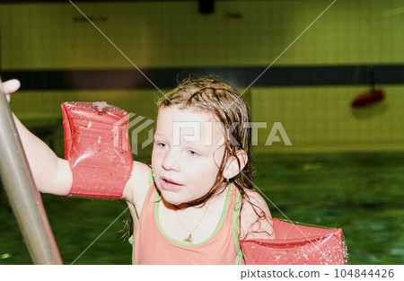 Small blond girl with armband floats in swimming pool. Child learns to swimm. Girl goes up on ladder in public pool. 104844426