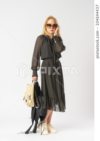 A woman in sunglasses and a casual dress holds a jacket and backpack in her hands 104844437