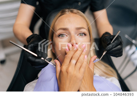 Closeup portrait of scared blonde young woman closing mouth by fear in dental clinic, afraid of treatment. Cropped shot of unrecognizable female doctor holding dental tool. Concept of dental treatment Closeup portrait of scared blonde young woman closing mouth by fear in dental clinic, afraid of treatment. Cropped shot of unrecognizable female doctor holding dental tool. Concept of dental treatment 104846393