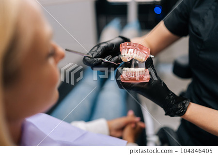 Close-up cropped shot of unrecognizable orthodontist in rubber gloves showing artificial human jaws with dental braces for teeth correction to female patient, visiting doctor orthodontic issues. Close-up cropped shot of unrecognizable orthodontist in rubber gloves showing artificial human jaws with dental braces for teeth correction to female patient, visiting doctor orthodontic issues. 104846405