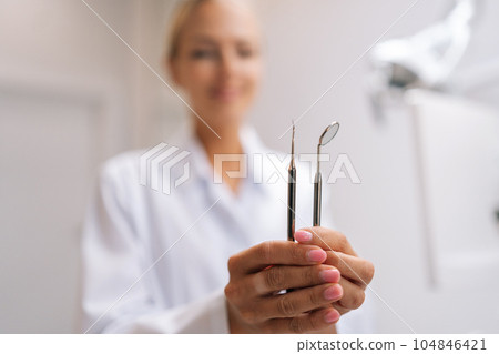 Closeup selective focus shot of female dental doctor holding working tools looking at camera in dentistry clinic with modern white interior. Concept of stomatology, dentistry and orthodontics. 104846421