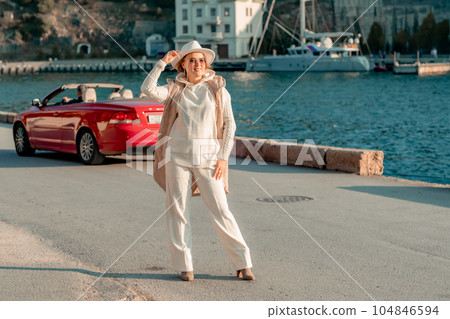 Portrait of a blonde convertible. Fashionable attractive woman with blond hair in a white hat on the background of a red car and the sea. Portrait of a blonde convertible. Fashionable attractive woman with blond hair in a white hat on the background of a red car and the sea. 104846594