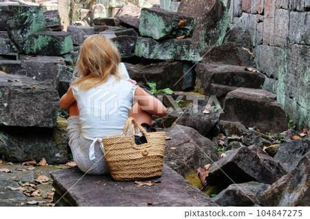 A young blonde European girl is sitting on a stone among medieval ruins and reading a book. A young blonde European girl is sitting on a stone among medieval ruins and reading a book. 104847275