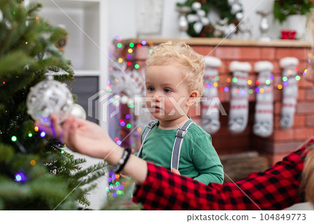 A little boy stands ip by the Christmas tree and watches the ornaments hanging on the branches. A little boy stands ip by the Christmas tree and watches the ornaments hanging on the branches. 104849773