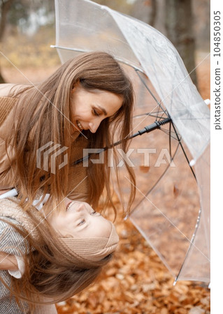 Young woman and little girl standing in autumn forest under transparent umbrella. Little girl looking at her mother. Girl wearing fashion grey jacket and woman brown one. Young woman and little girl standing in autumn forest under transparent umbrella. Little girl looking at her mother. Girl wearing fashion grey jacket and woman brown one. 104850305