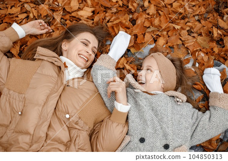 Young woman and little girl in autumn forest. Woman and her daughter lay on a fallen leaves and smilling. Girl wearing fashion grey jacket and woman brown one. 104850313