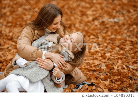 Young woman and little girl in autumn forest. Woman and her daughter sitting on a fallen leaves and hugging. Girl wearing fashion grey jacket and woman brown one. 104850315