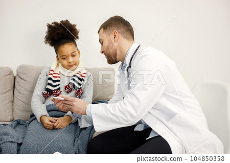 Male doctor asking sick girl about her symptoms. Girl has fever and overspread blanket. Doctor showing her a thermometr. Male doctor asking sick girl about her symptoms. Girl has fever and overspread blanket. Doctor showing her a thermometr. 104850358