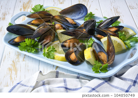 Steamed Mussels in shells in a blue plate with herbs and lemon. White wooden background. 104850749