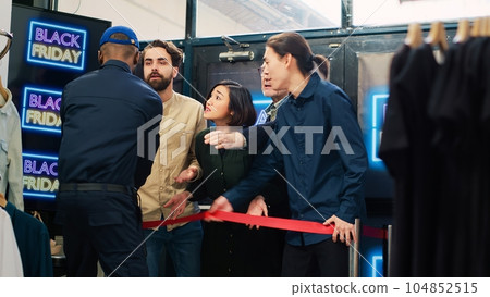 Customers pushing red tape and entering clothing store, looking to buy clothes on black friday sale. Security guard working under pressure with crowd of obsessed aggressive shoppers. Handheld shot. Customers pushing red tape and entering clothing store, looking to buy clothes on black friday sale. Security guard working under pressure with crowd of obsessed aggressive shoppers. Handheld shot. 104852515