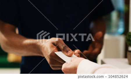 Male tourist receiving room key card for accommodation access after check in at front desk lobby. Seaside resort tourist registering at reception with hotel employee. Close up. Handheld shot. Male tourist receiving room key card for accommodation access after check in at front desk lobby. Seaside resort tourist registering at reception with hotel employee. Close up. Handheld shot. 104852624