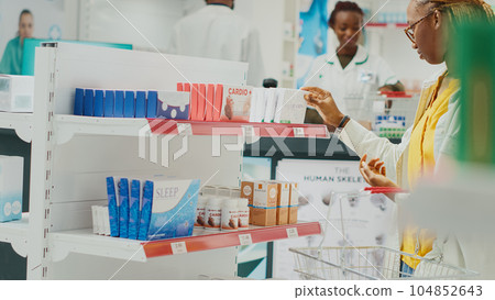 African american client looking at drugs on shelves, buying medicaments and healthcare products from drugstore. Young customer taking pills or vitamins from racks. Handheld shot. 104852643
