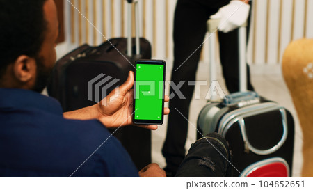 Travellers using greenscreen on phone, sitting in lounge area at hotel and looking at mockup copyspace. Couple waiting for registration procedure, holding smartphone with chromakey display. 104852651