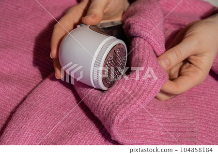 Woman using Anti-pilling razor at home. Device for shaving clothes. Anti-Plush fabric Shaver. Electric portable sweater pill defuzzer Lint remover from pink acrylic or wool sweater. Slow fashion Woman using Anti-pilling razor at home. Device for shaving clothes. Anti-Plush fabric Shaver. Electric portable sweater pill defuzzer Lint remover from pink acrylic or wool sweater. Slow fashion 104858184