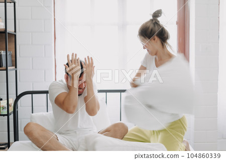 Funny couple angry wife hit husband with pillow in white bedroom. Funny couple angry wife hit husband with pillow in white bedroom. 104860339