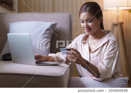 Pretty asian woman shopping online with credit card and laptop while sitting on sofa at home Pretty asian woman shopping online with credit card and laptop while sitting on sofa at home 104860696