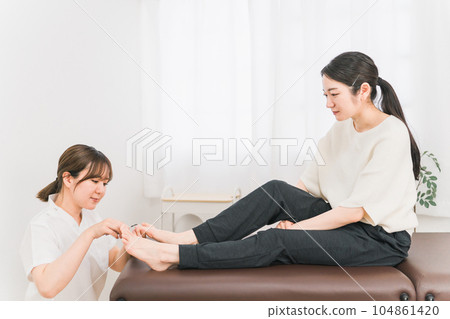 一名婦女和一名身穿白大衣的患者正在治療向內生長的腳趾甲和指甲 一名婦女和一名身穿白大衣的患者正在治療向內生長的腳趾甲和指甲 104861420