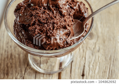 A transparent bowl with chocolate mousse - traditional French dessert and a teaspoon on a wooden table close-up 104862692