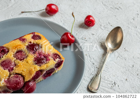 A slice of clafoutis french cherry pie in a gray plate and a spoon on the gray table, top view 104862722