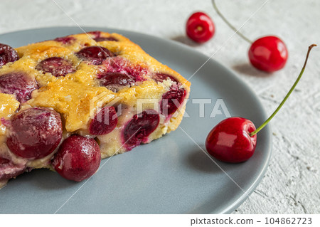 A slice of clafoutis cherry pie in a gray plate and fresh cherries on the gray table close-up 104862723