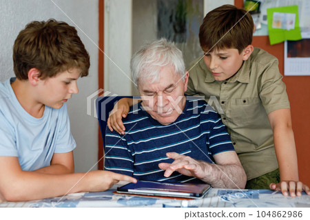 Two Preteen Boys Teaching Grandfather How to Use Internet Safely. Teenage Brothers, School Children with Digital Tablet Playing with Granddad, Senior Man at Home. 104862986