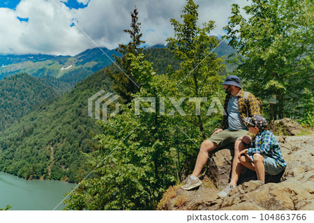 Hiking traveller caucasian bearded man with backpack sitting on rocks with son enjoying beautiful view of turquois blue Lake Ritsa in Abkhazia 104863766