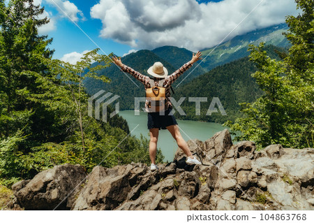 Hiking traveller woman with backpack hands up enjoying beautiful view of turquois blue Lake Ritsa in Abkhazia 104863768
