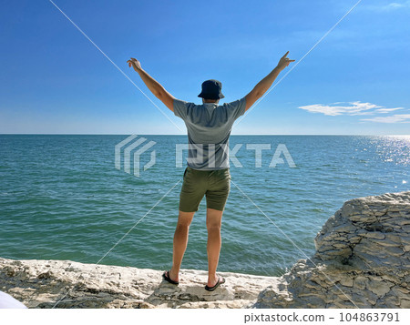 Traveller man with hands up enjoying beautiful sea view from rocky shore Traveller man with hands up enjoying beautiful sea view from rocky shore 104863791