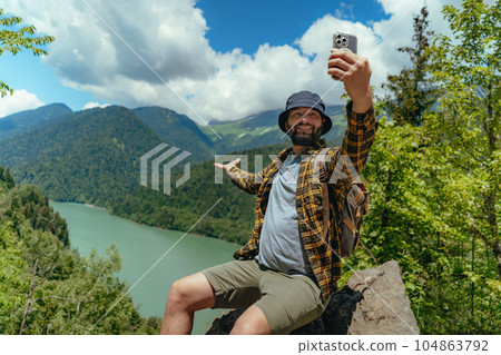 Hiking traveller handsome caucasian bearded man with backpack taking selfie against view of Lake Ritsa in Abkhazia Hiking traveller handsome caucasian bearded man with backpack taking selfie against view of Lake Ritsa in Abkhazia 104863792