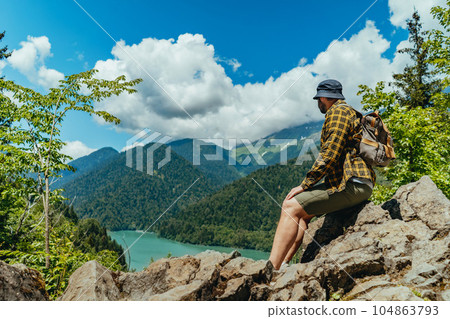 Hiking traveller caucasian bearded man with backpack sitting on rocks enjoying beautiful view of turquois blue Lake Ritsa in Abkhazia Hiking traveller caucasian bearded man with backpack sitting on rocks enjoying beautiful view of turquois blue Lake Ritsa in Abkhazia 104863793
