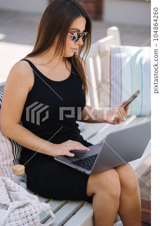 Beautiful young woman working outdoors and using phone and laptop at the same time Beautiful young woman working outdoors and using phone and laptop at the same time 104864260